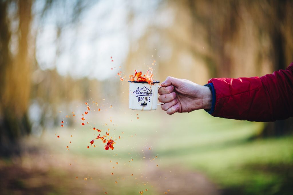 A hand tosses coffee from an enamel camp mug in a sunlit park